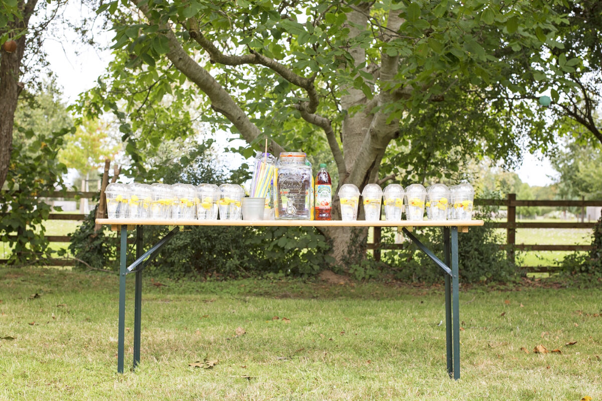 Image shows a party table under a tree in the shade. Atop of it are rows and rows of clear plastic cups with dome shaped lids, each filled with water and a yellow rubber duck.  There is a tin of coloured straws, squash bottles and a large mason drinks dispenser filled with water.