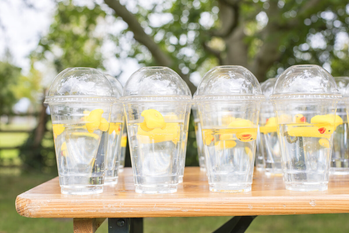 Image shows a table covered in rows of clear domed water cups with lids, filled with water. Each cup has a miniature yellow rubber duck floating in it.