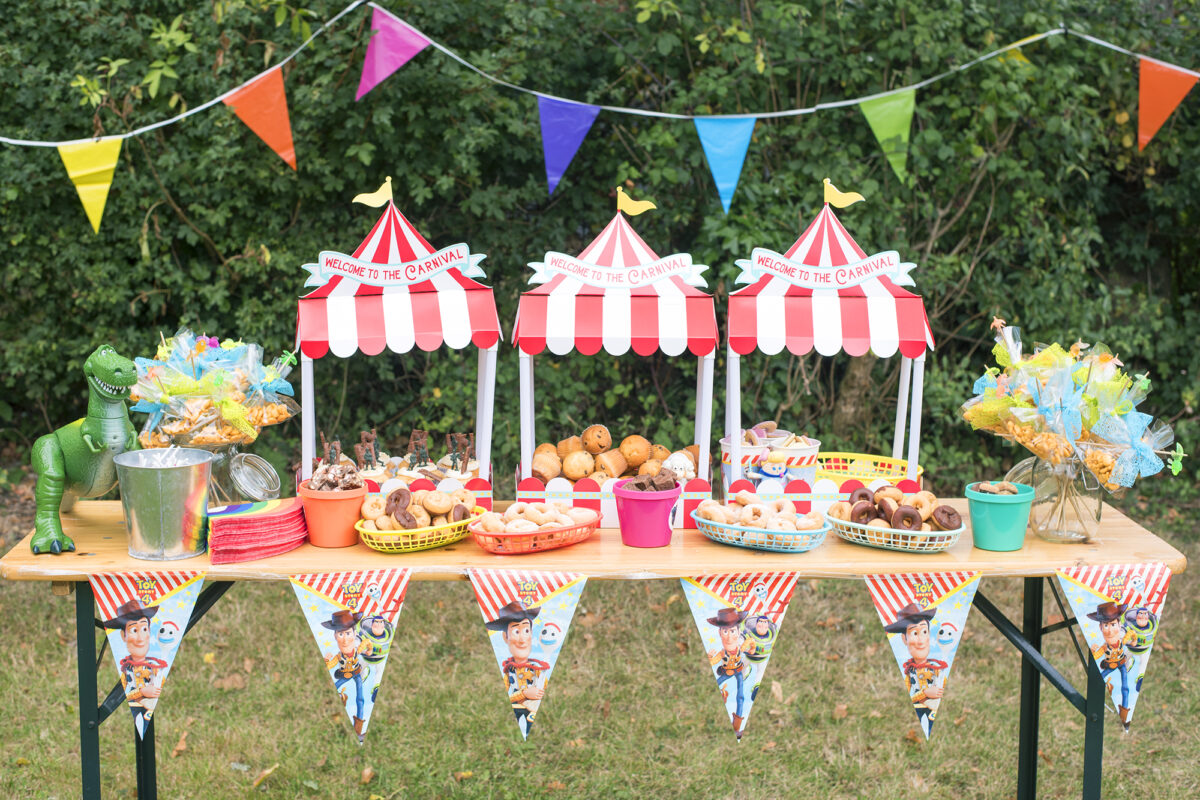 Image Shows a Toy Story party themed table outside in a garden.  There are three cardboard food stands in the shape of red and white big tops, which read welcome to the carnival. On the corner of the table is Disney's Rex from Toy Story toy and there are rainbow napkins, toy story bunting and the table is covered in Toy Story Army soldier cupcakes, mini donuts in different flavours, mini muffins, crackers and biscuits.