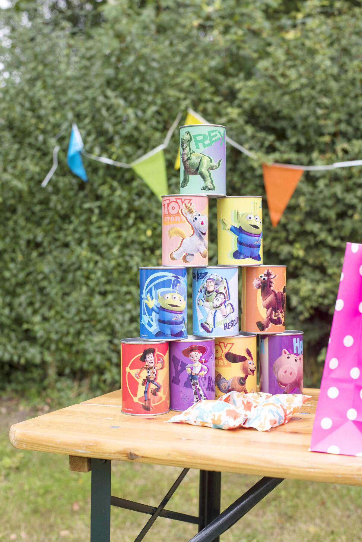 Image shows a wooden party table outside with a Toy Story themed set of knock down cans and bean bags in bright colours.  The table is outdoors in a field.
