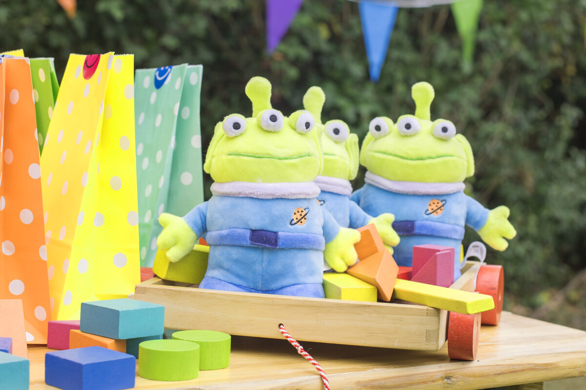 Image shows three plush Toy Story Alien toys from shopdisneyuk on top of a party table, sat with brightly coloured wooden blocks and party bags.