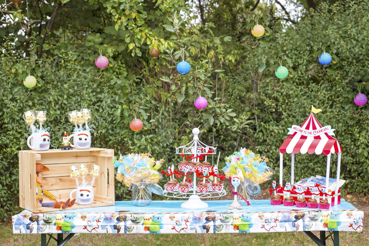 Image shows a party table outdoors in a field with a backdrop of hedges covered with coloured paper lanterns.  On the party table are carnival decorations, and Woody the Cowboy from Disney's Toy Story. There are mugs in the shape of Forky from Toy Story 4 which are filled with cake pops in the shape of Ducky from the film.  A Forky toy sits on the table next to a miniature food tent which says welcome to the carnival. There is a carousel for cupcakes filled with forky cupcakes, and bags of pepperidge farm goldfish crackers on fishing poles  in glass jars with little plastic goldfish sat on top. 