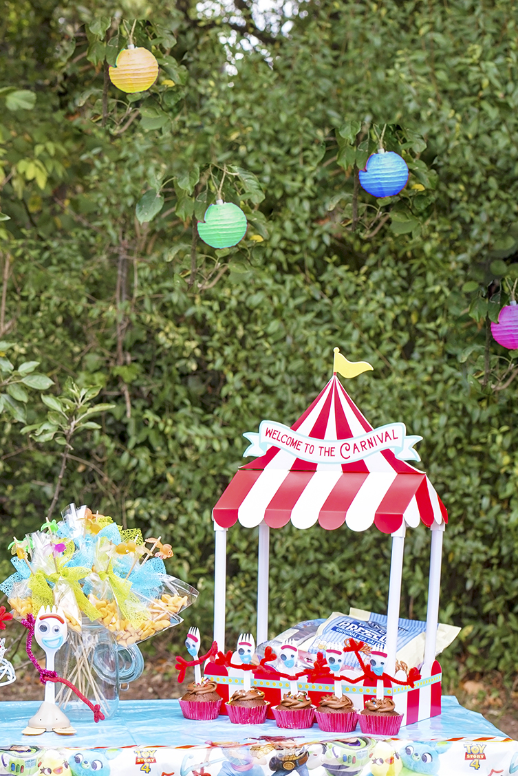 Image shows a party table set in a carnival theme with Disney's Toy Story Forky figure sitting next to a red and white striped carnival big top table decoration filled with pretzels and forky cupcakes lined along the front.