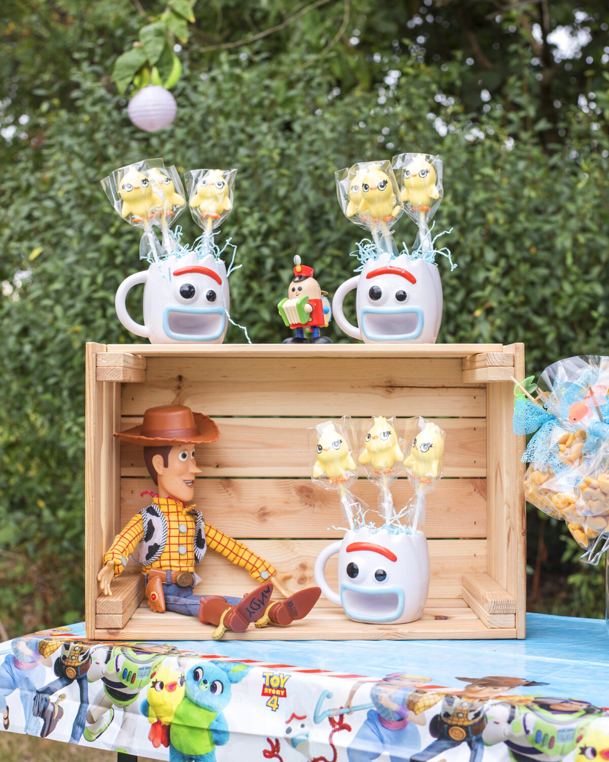 Image shows a party table with an upturned wooden ikea crate. Inside the crate sits a toy of Woody the Cowboy from Disney's Toy Story, and on top are three Forky shaped mugs filled with Ducky shaped cake pops. The original toy story pixar tinker toy is on top of the crate.