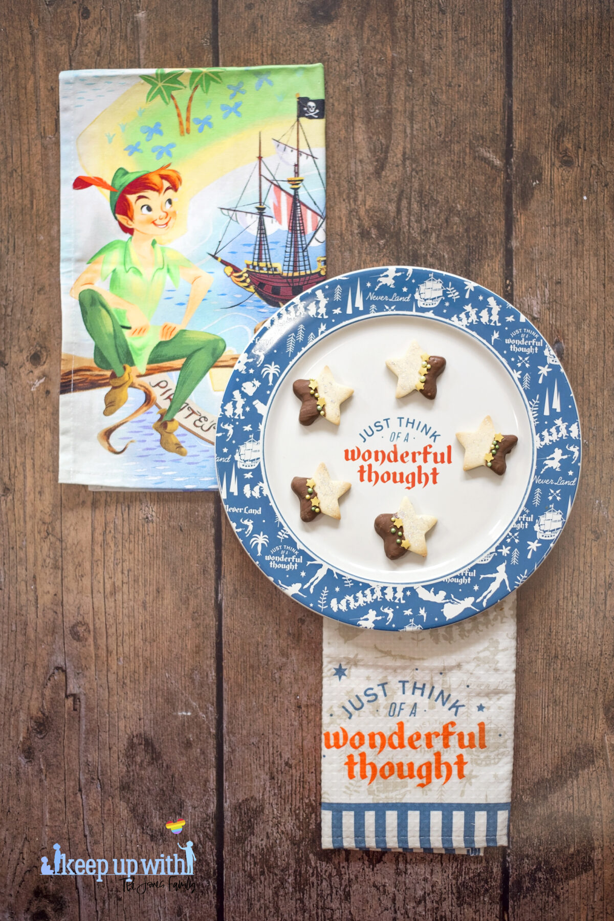 Image shows a wooden tabletop with ShopDisneyUK's Peter Pan tea towel set and a Peter Pan dinner plate reading "just think of a wonderful thought" on it.  On the Disney plate are small star shaped biscuits half of each Peter Pan Second Star to the Right Biscuit is dipped in chocolate and  the other is sprinkled with silver edible glitter.  The dividing line is decorated with yellow star sprinkles and green pearl shaped sprinkles. Image by Keep Up With The Jones Family