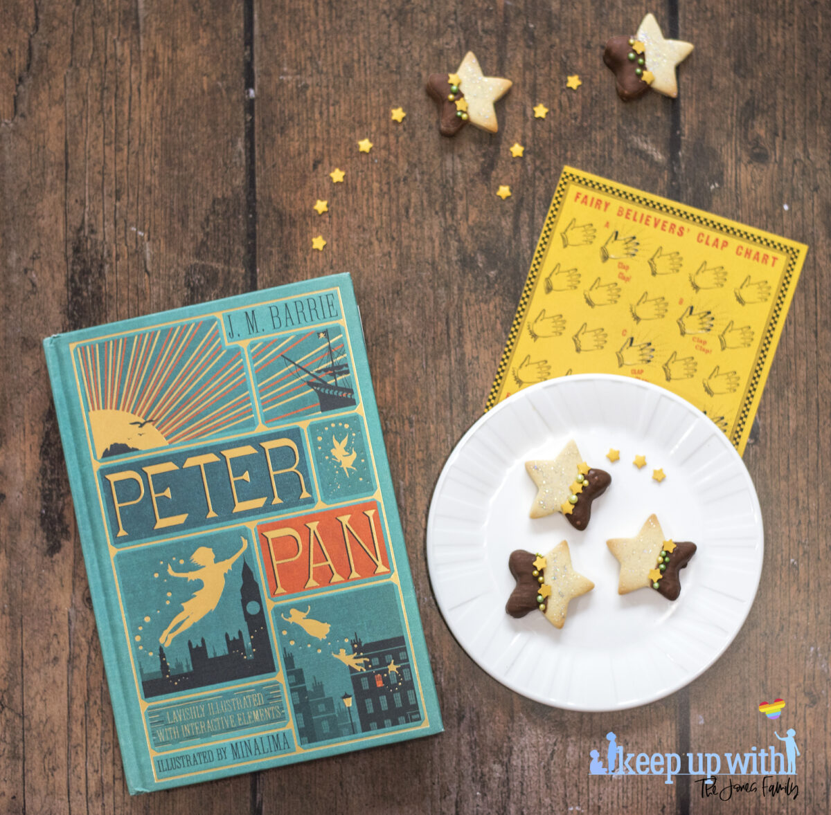 Image shows a wooden tabletop with a copy of J M Barrie's Peter Pan, a small white Vera Wang Wedgewood plate and a fairy clapping chart,  On the table are small star shaped biscuits. Half of each Peter Pan Second Star to the Right Biscuit is dipped in chocolate and  the other is sprinkled with silver edible glitter.  The dividing line is decorated with yellow star sprinkles and green pearl shaped sprinkles. Image by Keep Up With The Jones Family