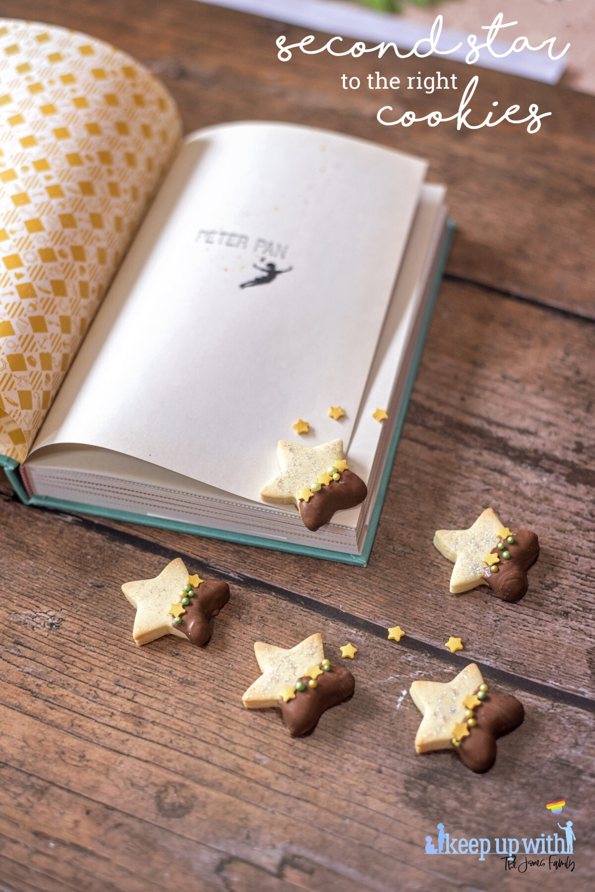Image shows a wooden tabletop with a copy of J M Barrie's Peter Pan open. On the book pages and the table are small star shaped biscuits. Half of each Peter Pan Second Star to the Right biscuit is dipped in chocolate and  the other is sprinkled with silver edible glitter.  The dividing line is decorated with yellow star sprinkles and green pearl shaped sprinkles. Image by Keep Up With The Jones Family