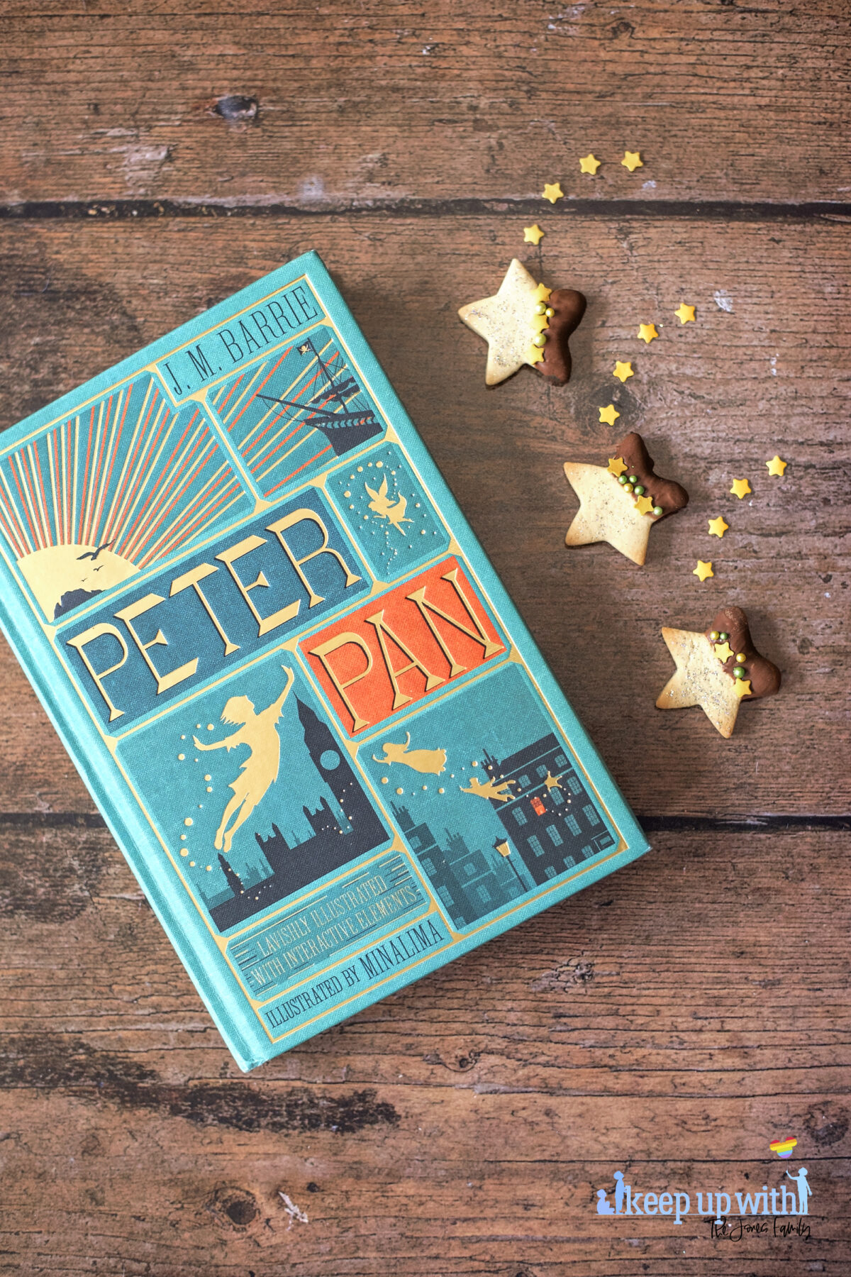Image shows a wooden tabletop with a copy of J M Barrie's Peter Pan. On the book pages and the table are small star shaped biscuits. Half of each biscuit is dipped in chocolate and  the other is sprinkled with silver edible glitter.  The dividing line is decorated with yellow star sprinkles and green pearl shaped sprinkles. Image by Keep Up With The Jones Family