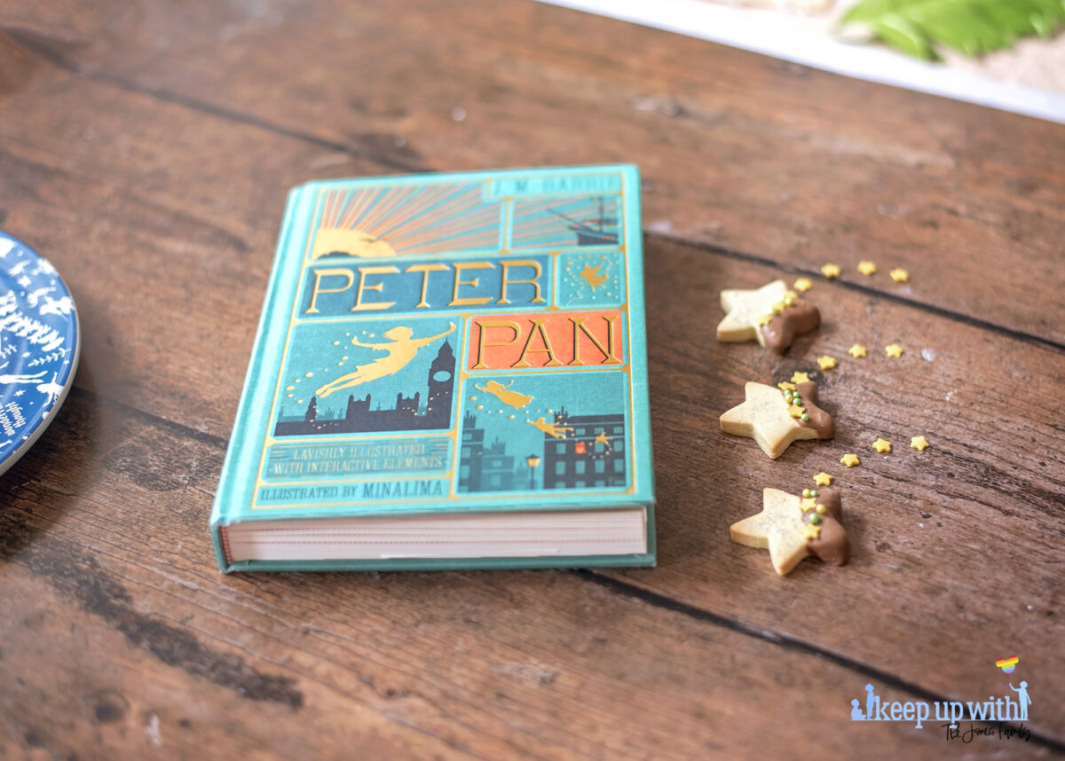 Image shows a wooden tabletop with a copy of J M Barrie's Peter Pan. On the book pages and the table are small star shaped biscuits. Half of each Peter Pan Second Star to the Right Biscuit is dipped in chocolate and  the other is sprinkled with silver edible glitter.  The dividing line is decorated with yellow star sprinkles and green pearl shaped sprinkles. Image by Keep Up With The Jones Family