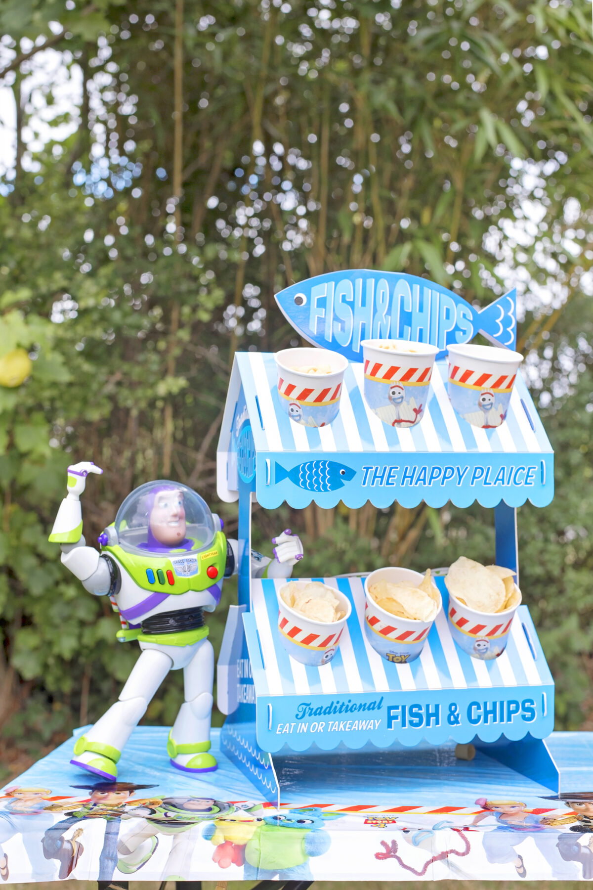 Image shows a party table outdoors with Disney's Buzz Lightyear toy stood on top of it, next to a novelty fish and chip stand made from cardboard.  