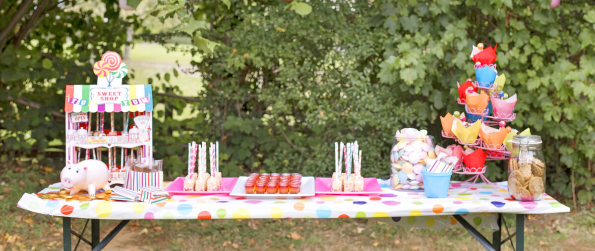 Image shows a party table set with rainbow jellies, brightly coloured chocolate cupcakes with lollipops, a jar of chocolate chip cookies, rice krispie lollipops covered in rainbow sprinkles, and a little sweet shop stand filled with chocolate dipped marshmallows. A Hamm ceramic money box from Disney's Toy Story 4 is on the table sat on top of a mountain of gold chocolate coins.  The table is outdoors at the party in a field with hedges behind it.
