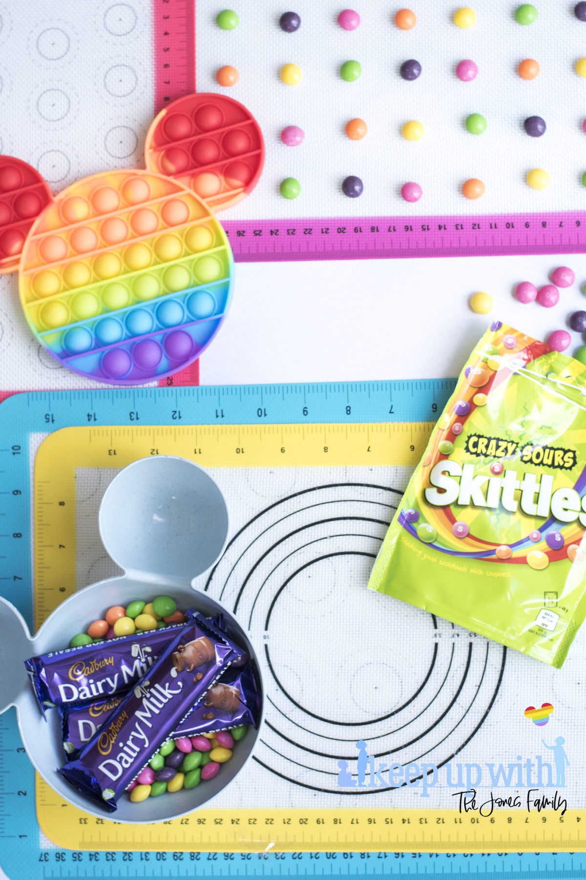 Image shows a rainbow coloured pop-it toy on top of rainbow coloured silicone baking trays. There is a blue mickey mouse shaped bowl full with skittles sweets and the packet by the side.  A Disney's Mickey Mouse Pop-It Chocolate Bar. Image by Keep Up With the Jones Family. 