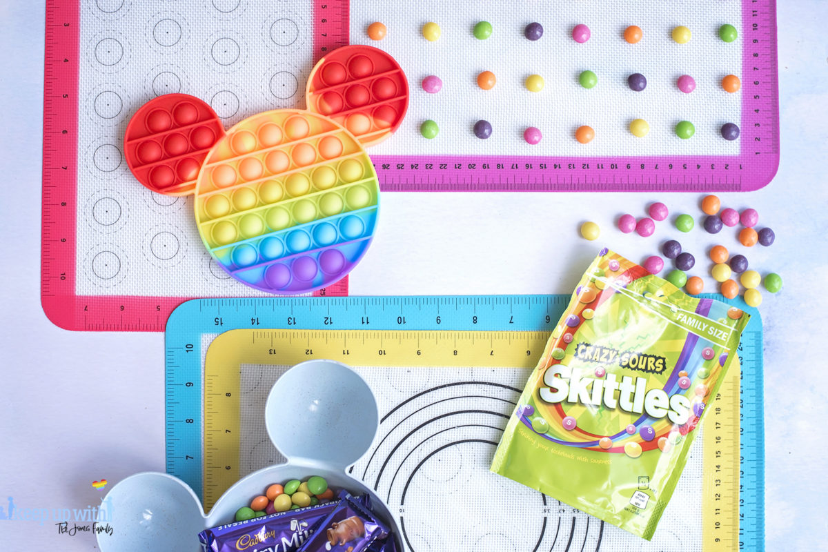 Image shows the ingredients for a Disney's Mickey Mouse Pop-It Chocolate Bar. IA rainbow coloured pop-it toy in the shape of Mickey Mouse, and some skittles sweets and candy.  Image by Keep Up With the Jones Family. 