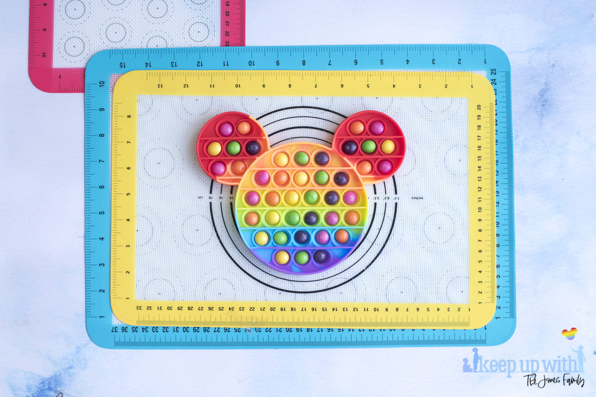 Image shows a Mickey Mouse shaped rainbow coloured silicone pop-it toy filled with skittles ready to make a Mickey Mouse Chocolate Pop-It bar. Image by Keep Up With the Jones Family. 