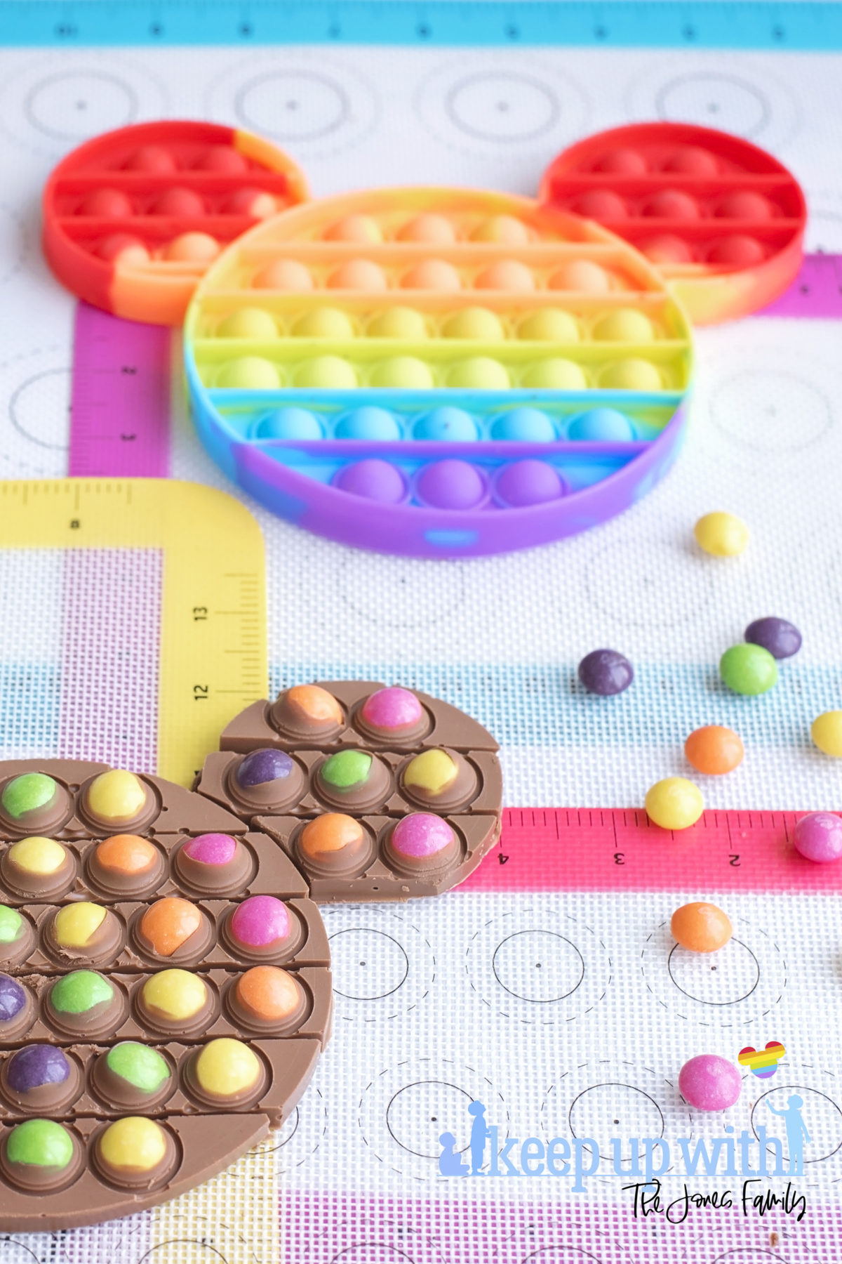 Image shows a rainbow Pop-It toy in the shape of Mickey Mouse on rainbow silicone baking trays, surrounded by skittles sweets and a chocolate bar dotted with the sweets. A Disney's Mickey Mouse Pop-It Chocolate Bar. Image by Keep Up With the Jones Family. 
