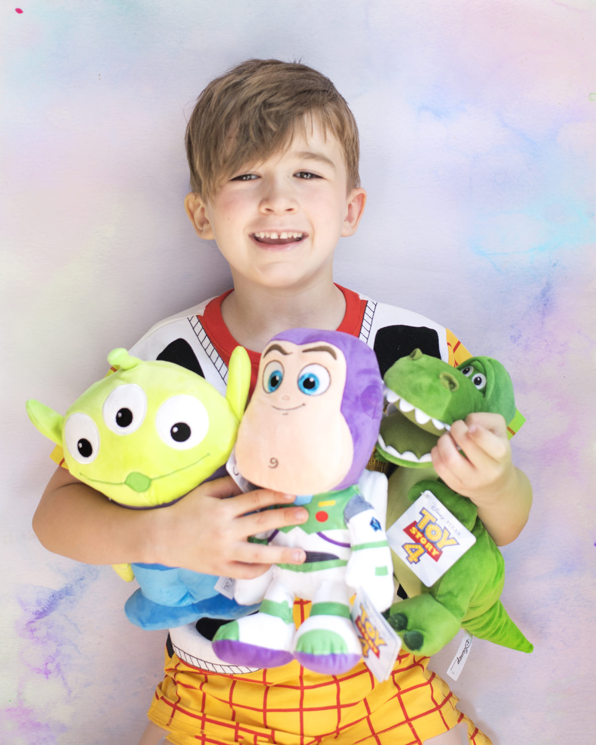 Image shows a young boy dressed in a Disney's Toy Story Woody t-shirt from NEXT, colding three Disney Pixar's Toy Story characters in his arms - Dinosaur, Rex, Buzz Lightyear and an Alien as plush toys from SImba Toys. Image by Keep Up With The Jones Family. 