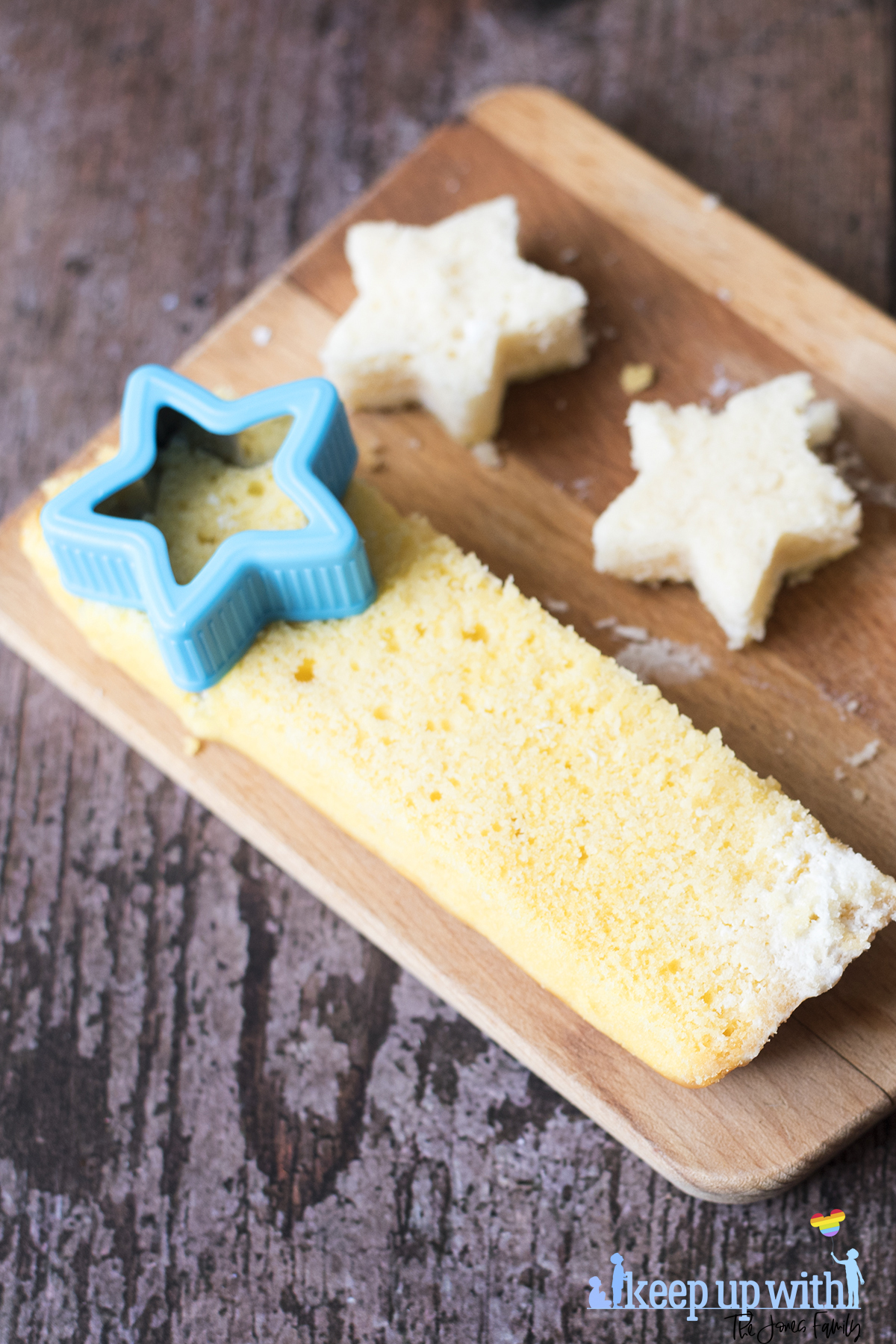 Image shows vanilla angel cake stars being cut from a slice of cake using a blue star cookie cutter, on top of a wooden chopping board, resting on a dark wooden tabletop. Image by Sara-Jayne Jones of Keep Up With The Jones Family.