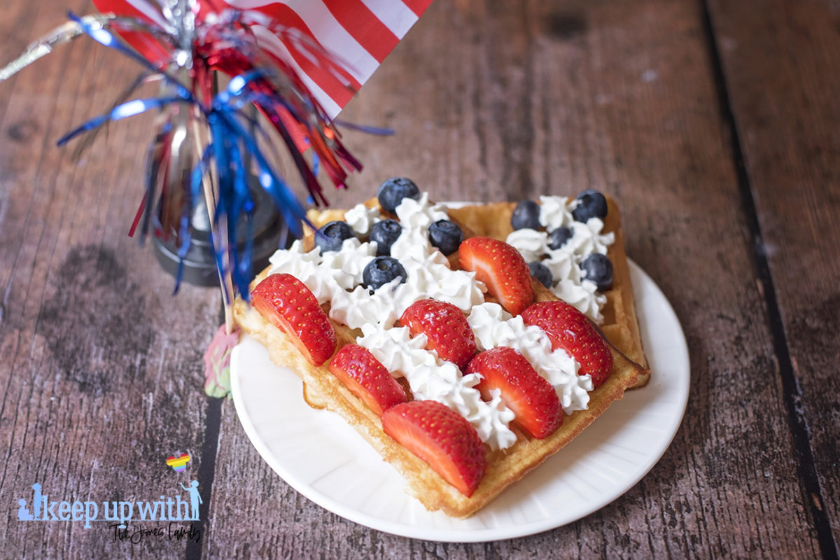 Image shows rectangular sweet breakfast waffles on a white vera wang plate. The waffles are decorated in the style of the American Flag. There are sliced strawberries for the red stripes, blueberries for the space around the stars, and cream piped as the white stars and stripes. The plate is set on a dark hardwood background and there is a small American Flag next to it and a red, silver and blue set of firework decorations. Image by Sara-Jayne Jones, Keep Up With The Jones Family.