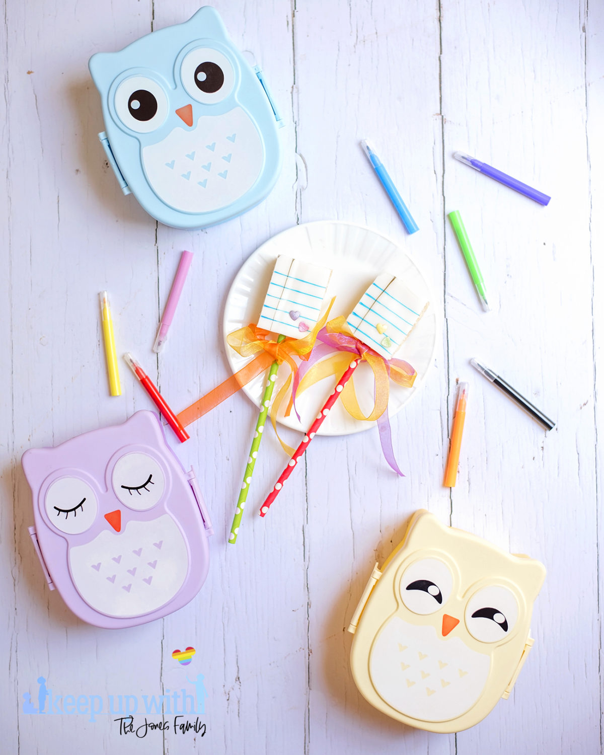 Image shows Back to School Rice Krispie Lollipop treats on a white Vera Wang for Wedgwood plate. There are three pastel coloured owl bento boxes in the background and a small tin bucket of Crayola crayons alongside it. Cake pens surround the plate on the white wooden background. Image by Sara-Jayne from Keep Up With The Jones Family.