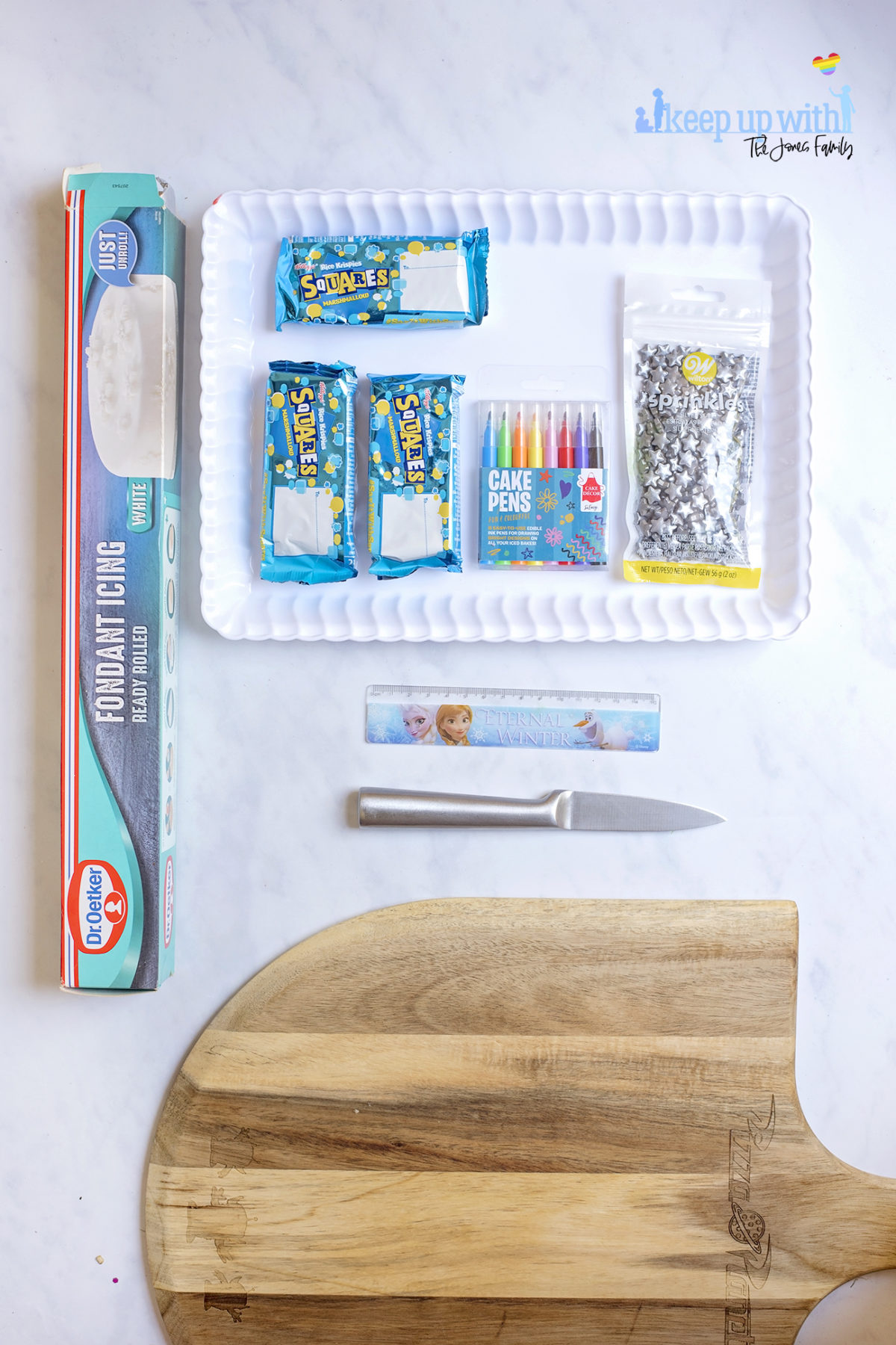 Image shows ingredients for Back to School Rice Krispie treats on a white scalloped tray, sitting on a white marble surface. There are Rice Krispies Squares Bars, sprinkles, white pre-rolled royal icing fondant, cake pens, a ruler, knife and shopping board. Image by Sara-Jayne from Keep Up With The Jones Family.
