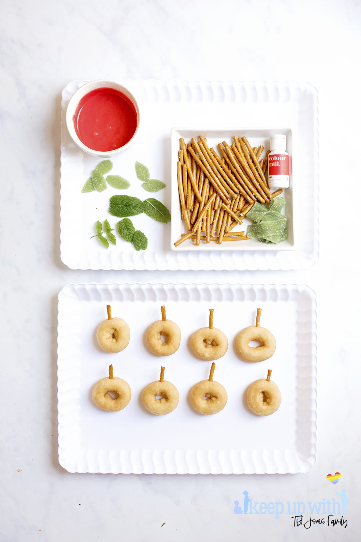 Image shows ingredients for Back to School Doughnuts on a white scalloped tray, sitting on a white marble surface. There are mini doughnuts, pretzel sticks, green fizzy belt sweets, mint leaves, red oil based food colouring and a bowl of icing sugar coloured red. Beneath the tray is a second tray with the mini donuts prepared with pretzel stalks in. Image by Sara-Jayne from Keep Up With The Jones Family.