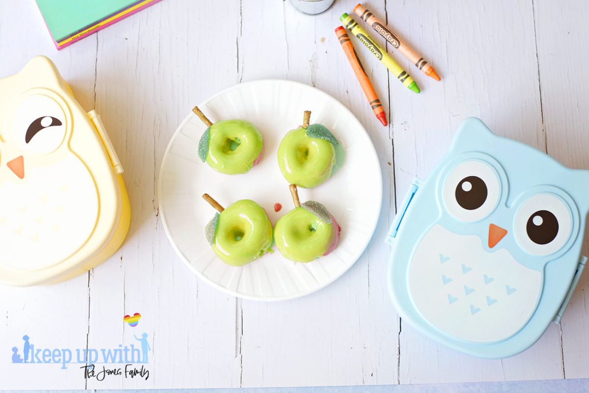 Image shows a flatlay of green apple shaped Back to School Doughnuts on a white Vera Wang plate, sitting on a white marble surface. Two owl shaped bento boxes and crayola crayons are in the photograph. Image by Sara-Jayne from Keep Up With The Jones Family.