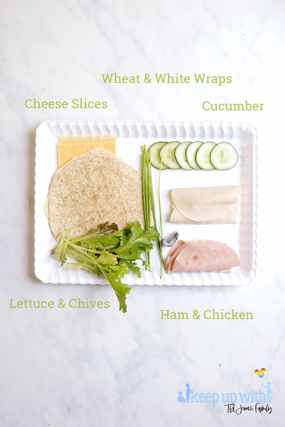 Image shows ingredients needed to make Book Sandwich Wraps. On a white scalloped tray, sat on a white marble top, are two white and wheat wraps, cucumber, ham and chicken slices, lettuce leaves, chives and cheese slices. Image by Sara-Jayne of Keep Up With The Jones Family.