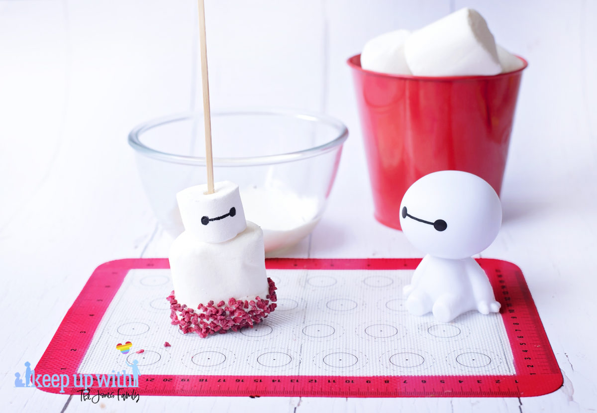 Image shows a plastic Baymax figure from Disney Pixar's Big Hero 6 sat on a white tabletop. In front of him is a fruity baymax marshmallow pop sat on a silicone baking mat. In the background is a glass bowl containing melted white candy drops and a red tin bucket of large marshmallows. Image by Keep Up With The Jones Family.