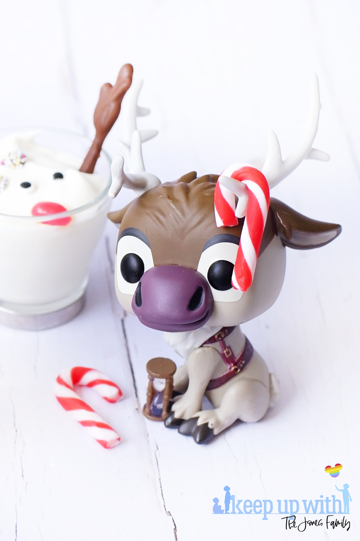 Image shows a Disney's Frozen Sven the Reindeer Funko Pop Vinyl decorated with mini red and white peppermint candy canes on his antlers. The figure is sitting next to a Melted Sven the Reindeer Yoghurts in glass cups with silver handles, on a white wooden tabletop. Image by Keep Up With The Jones Family.