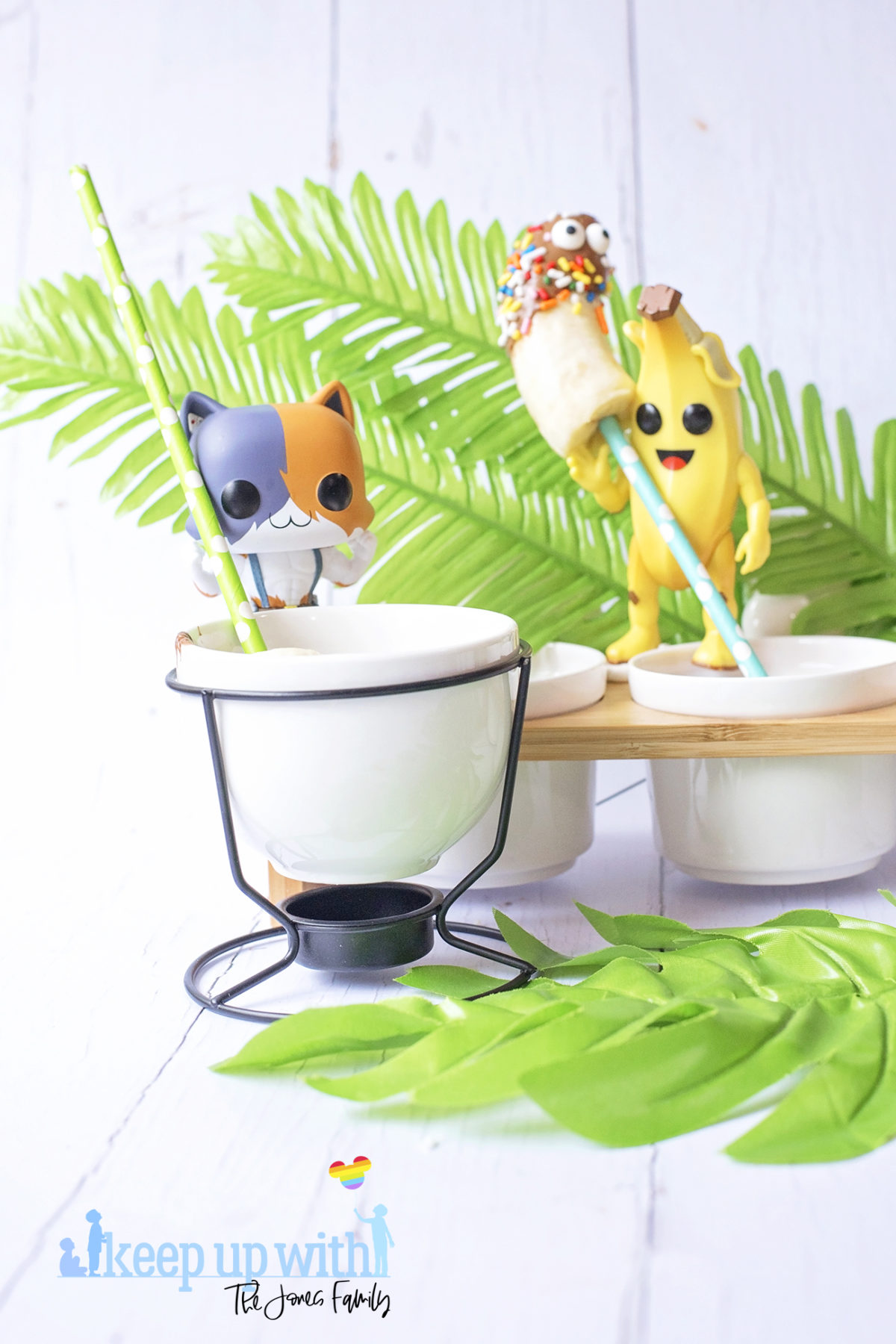 Image shows Peely’s Banana Fortnite Fondue Bar, fun food for families. A Funko Pop Vinyl Peely and Meowscles stand on a fondue bar stirring chocolate with bananas on sticks. Image by Sara-Jayne from Keep Up With The Jones Family.