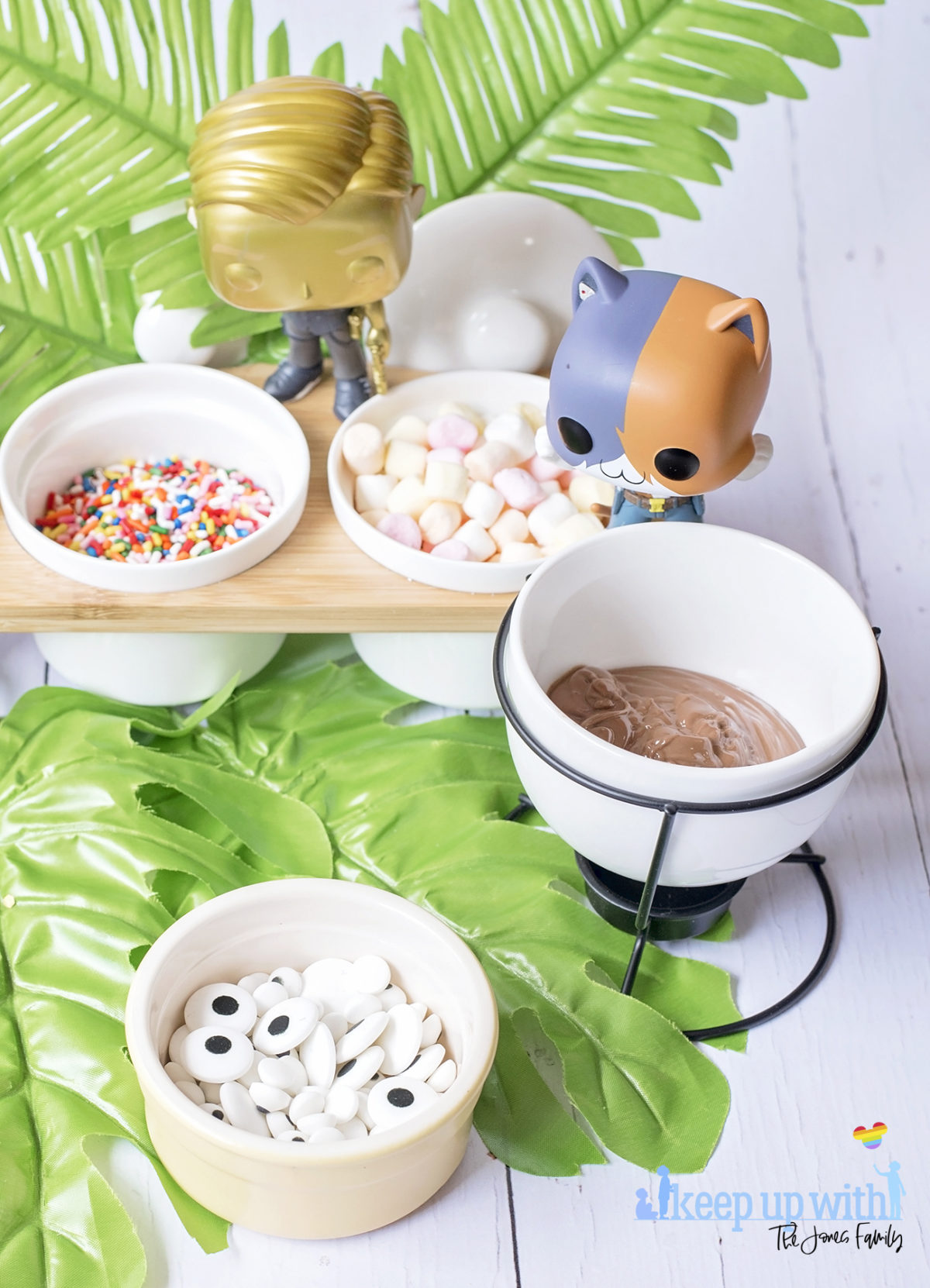 Image shows Peely’s Banana Fortnite Fondue Bar, fun food for families. A Funko Pop Vinyl Meowscles and Midas stand on a fondue bar next to dishes of fondue toppings including edible eyes, sprinkles, mini marshmallows and peanut butter drops. Image by Sara-Jayne from Keep Up With The Jones Family.