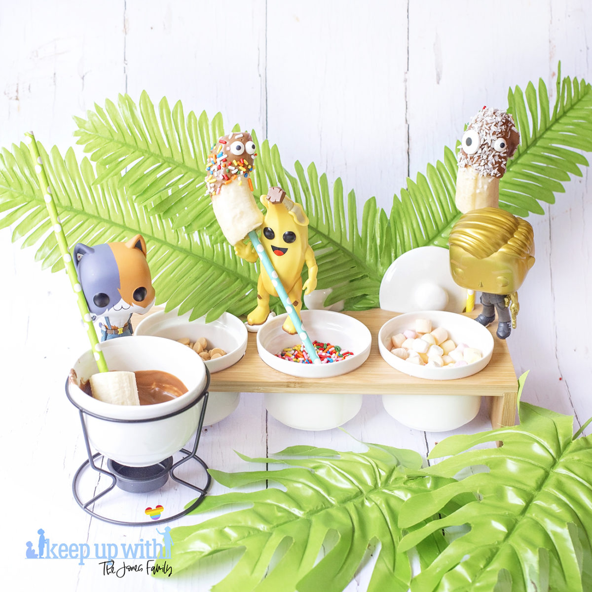 Image shows Peely’s Banana Fortnite Fondue Bar, fun food for families. A Funko Pop Vinyl Peely, Meowscles and Midas stand on a fondue bar next to bananas on spotted coloured straws, a chocolate fondue and various toppings including edible eyes, sprinkles, mini marshmallows and peanut butter drops. Image by Sara-Jayne from Keep Up With The Jones Family.