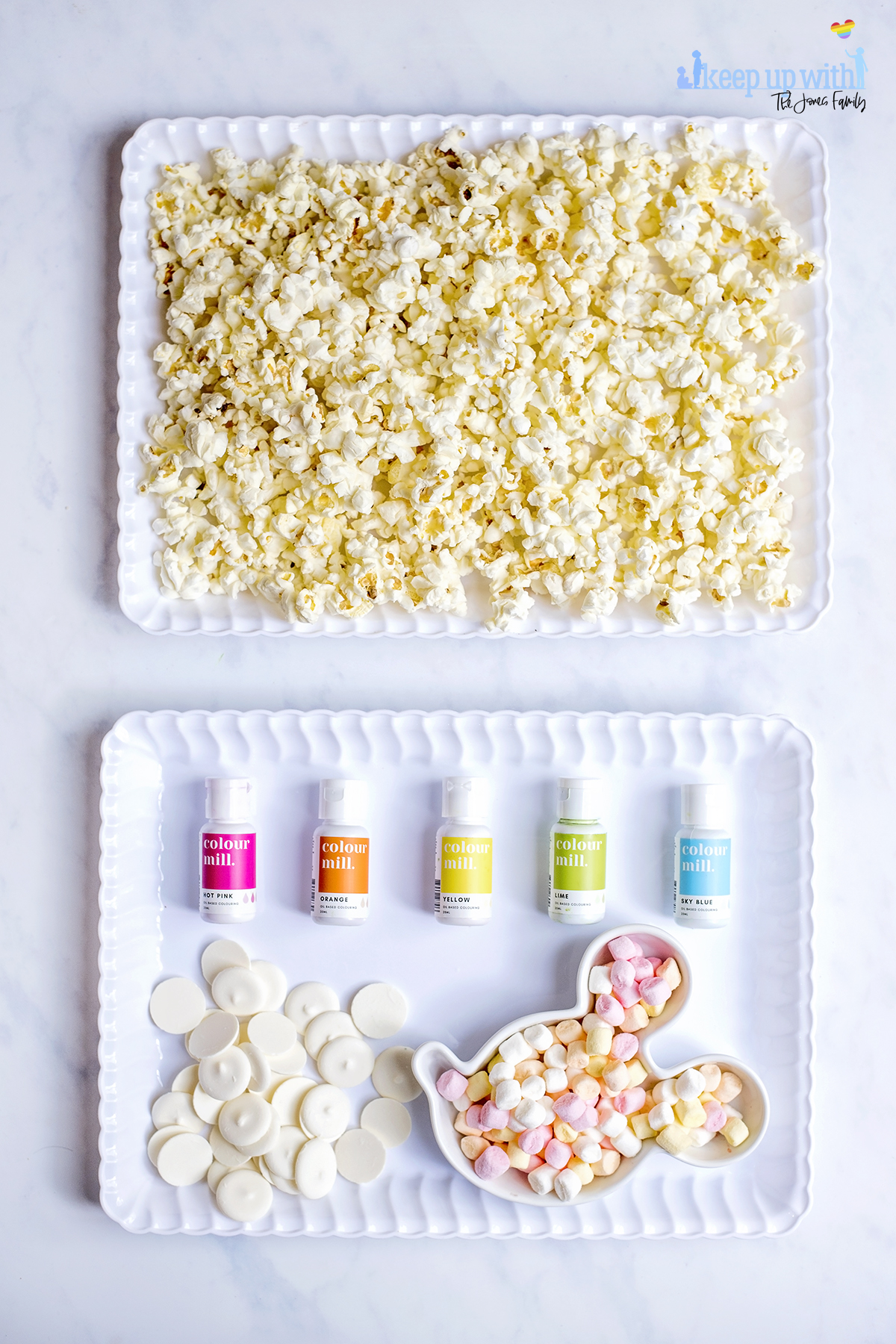 Image shows the ingredients used to make Mermaid Popcorn. Popcorn, spread evenly into a white scalloped tray, a pile of bright white candy melts, a mickey mouse shaped bowl of mini marshmallows and oil based food colourings from Colour Mill. Image by Sara-Jayne from keep Up With the Jones family.