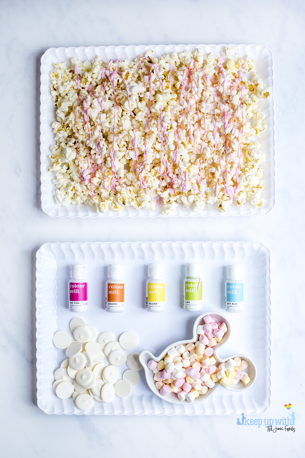 Image shows the ingredients used to make Mermaid Popcorn. Popcorn, spread evenly into a white scalloped tray, which has been coated in drizzled pink candy melts, a pile of bright white candy melts, a mickey mouse shaped bowl of mini marshmallows and oil based food colourings from Colour Mill. Image by Sara-Jayne from keep Up With the Jones family.
