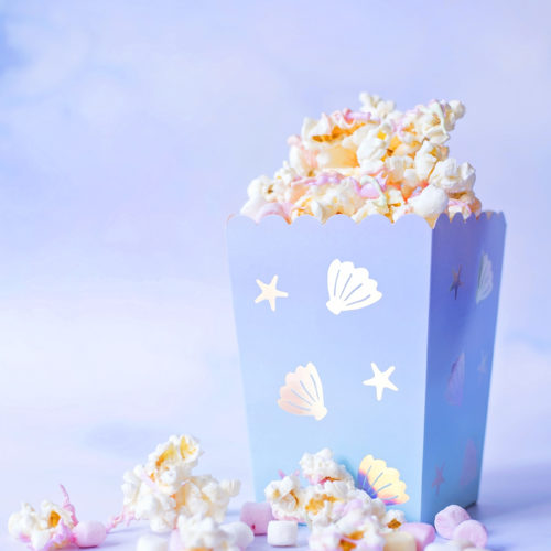 Image shows a carton of Mermaid Popcorn on a pale blue background. The popcorn is coated in pastel pink, orange and green candy melts, and there are mini pastel marshmallows and pearl sprinkles mixed in. The popcorn box is mermaid themed from Meri Meri. Image by Sara-Jayne from Keep Up With The Jones Family.