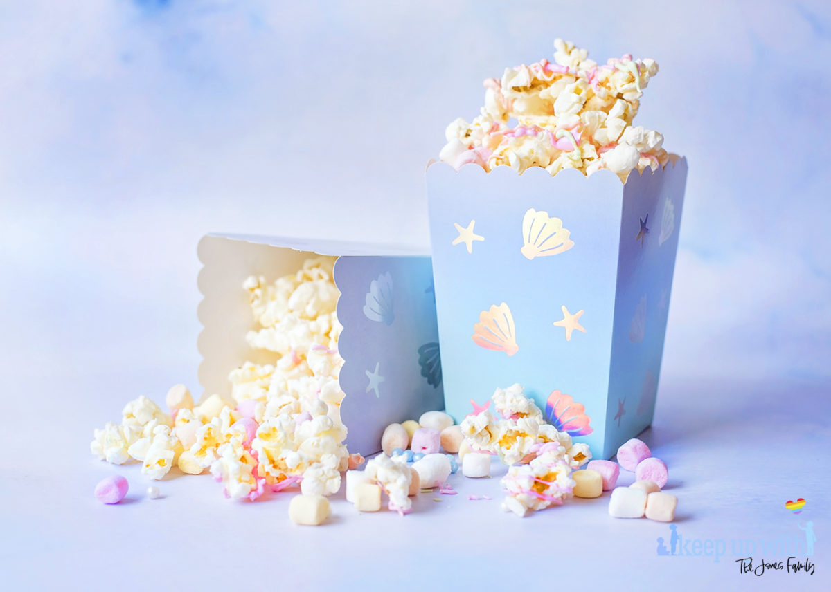 Image shows two cartons of Mermaid Popcorn on a pale blue background.  One is tipped over and popcorn is spilling out.  The popcorn is coated in pastel pink, orange and green candy melts, and there are mini pastel marshmallows and pearl sprinkles mixed in.  The popcorn box is mermaid themed from Meri Meri. Image by Sara-Jayne from Keep Up With The Jones Family.