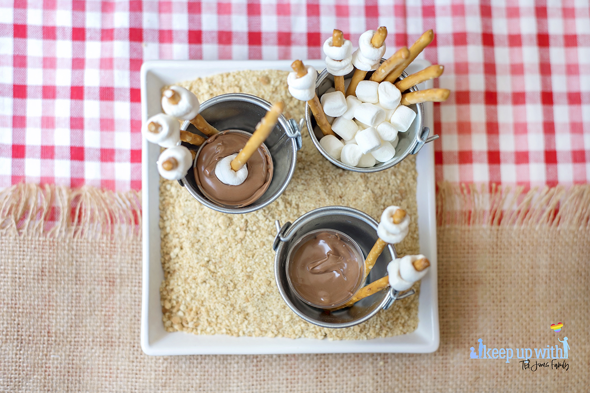 Image shows Mini Campfire Chocolate Fondue Buckets. Miniature galvanised steel buckets containing pretzel sticks with small marshmallows place on the ends like roasting marshmallow sticks, and a small plastic shot glass filled with melted chocolate. The buckets are placed on a white square plate filled with crushed digestive biscuit crumbs to look like sand. Image by Keep Up With The Jones Family.