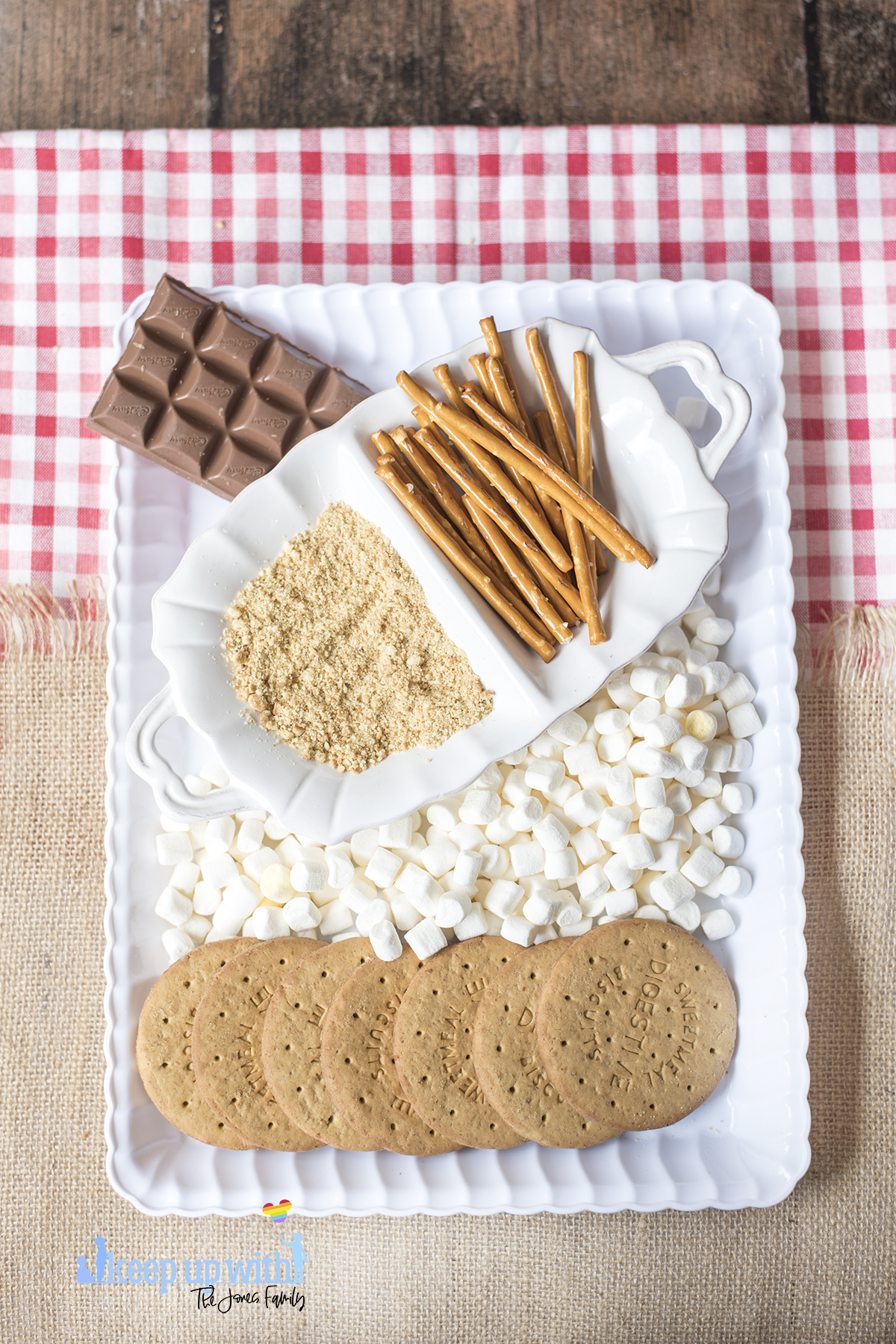 Image shows a white scalloped tray containing all of the ingredients to make Mini Campfire Chocolate Fondue Buckets. Mini marshmallows, digestive biscuits, pretzel sticks and chocolate. Image by Keep Up With The Jones Family.