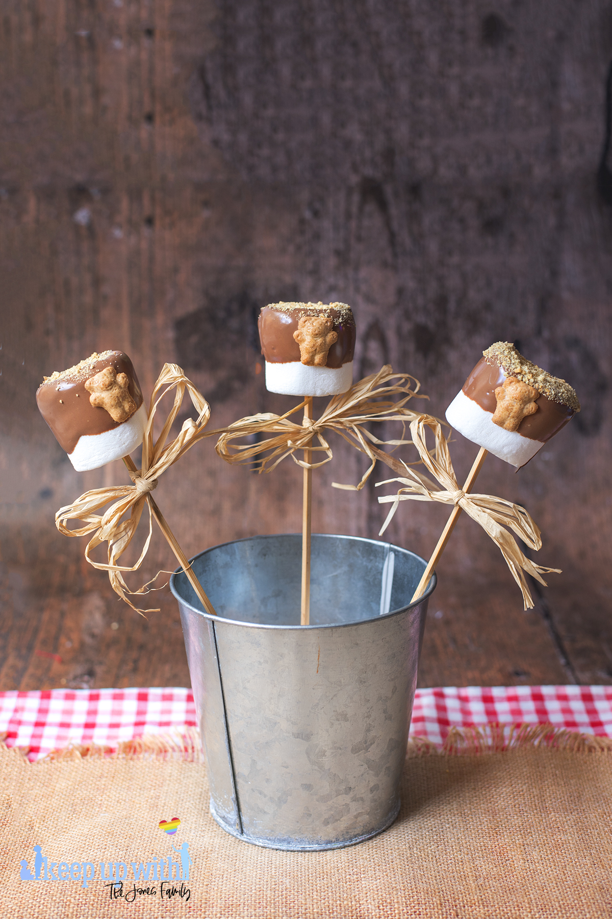 Image shows three teddy bear s'mores pops in a tin bucket on a dark wooden tabletop. Image by keep up with the jones family.