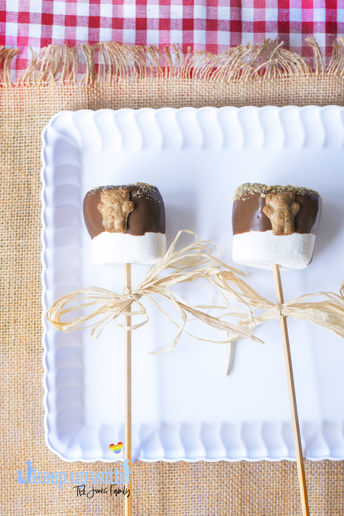 Image shows a white scalloped tray sat on top of a fringed jute tablecloth, which is on top of a red and white checked tablecloth. Inside the tray are two large teddy bear s'mores pops.